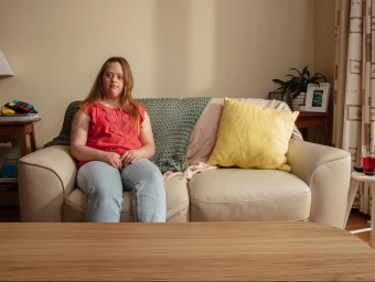 Young woman wearing a red top and light wash jeans seated on a light beige leather sofa in a living room.