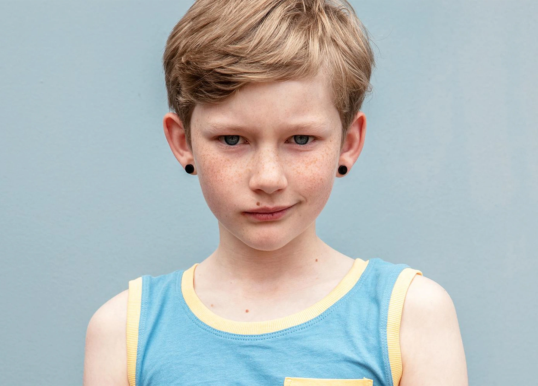 Portrait of a young child with short blonde hair and freckles wearing a blue tank top with yellow trim against a solid grey background