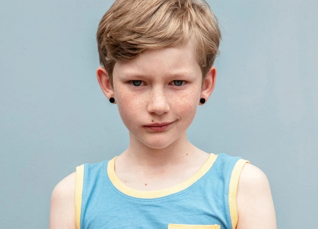 Portrait of a young child with short blonde hair and freckles wearing a blue tank top with yellow trim against a solid grey background