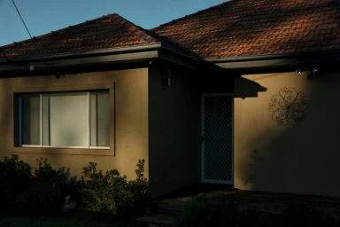 Exterior view of a single-story house with tan walls, dark tiled roof, a large window, and screen door photographed by Billy Plummer.