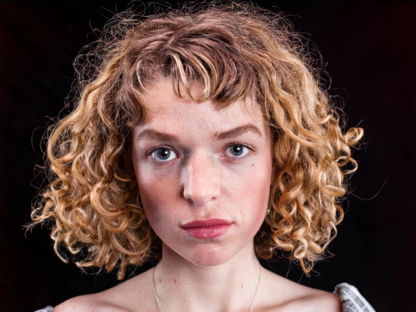 Close-up portrait of a woman with curly blonde hair and blue eyes against a dark background, photographed by Billy Plummer.