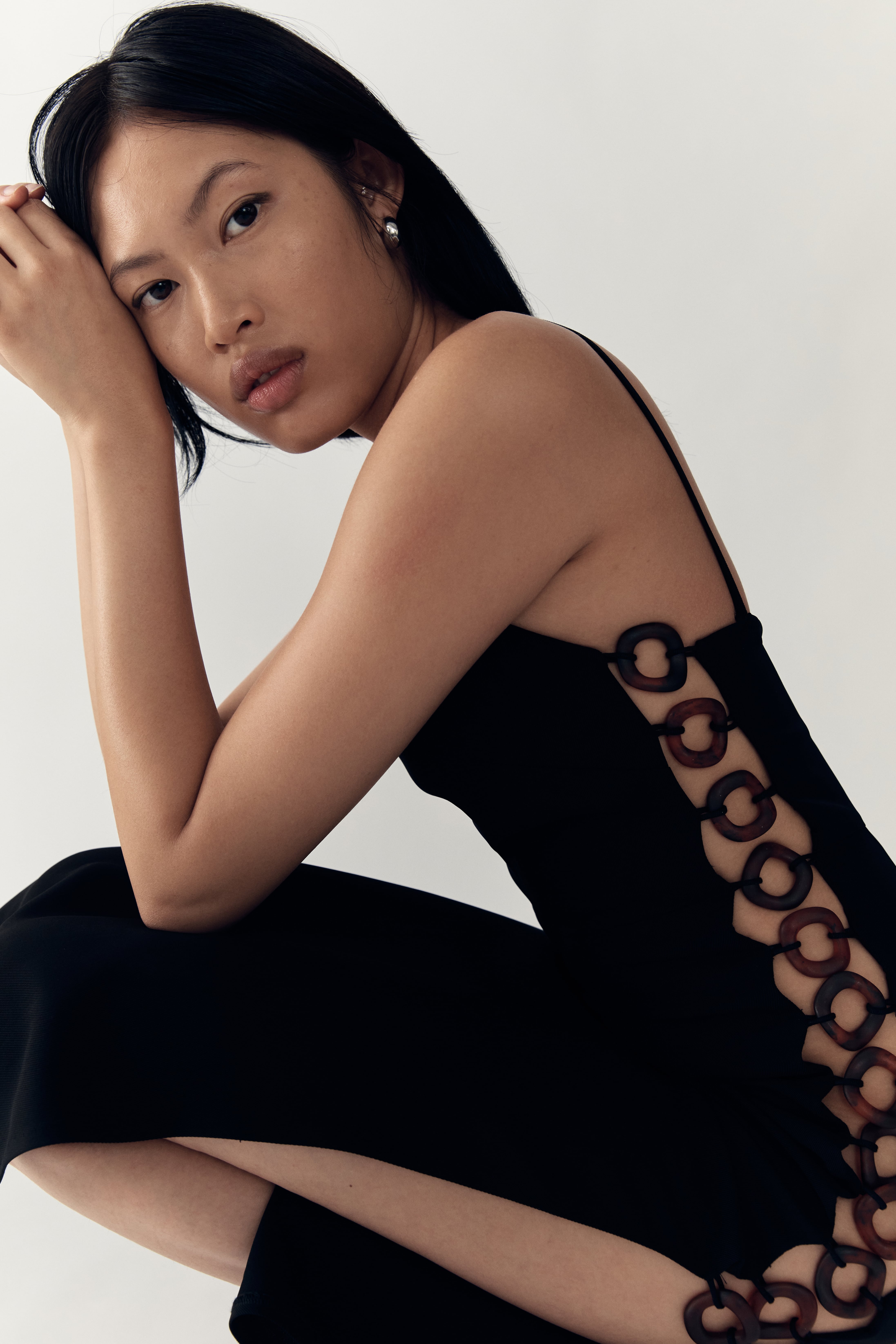 An Asian woman in a black dress with circular brown cutouts poses in a bright, clean photo studio with a minimalist fashion style.