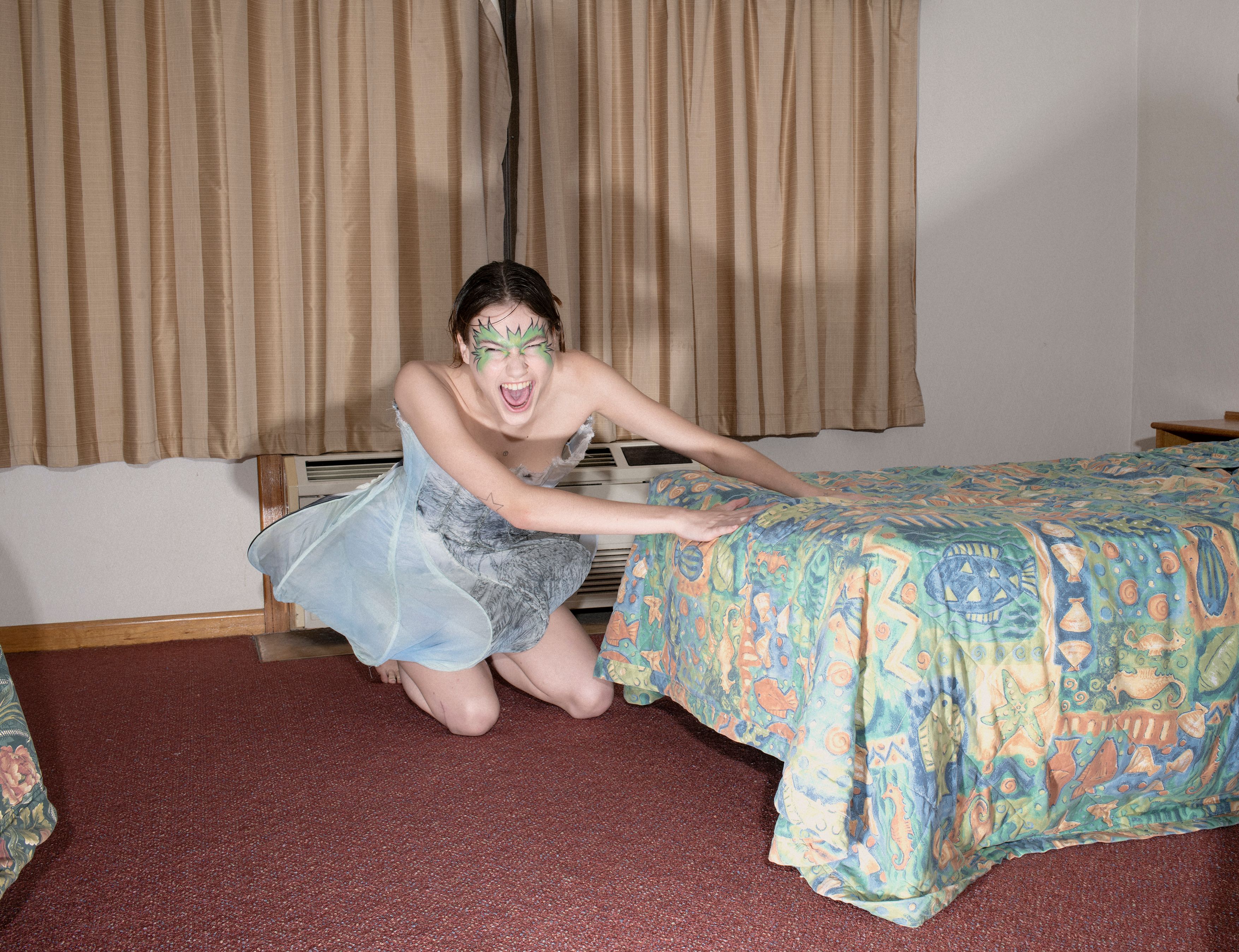 Dramatic image of a kneeling woman with face paint in a light blue dress, screaming in a motel room, capturing raw emotion and vulnerability.