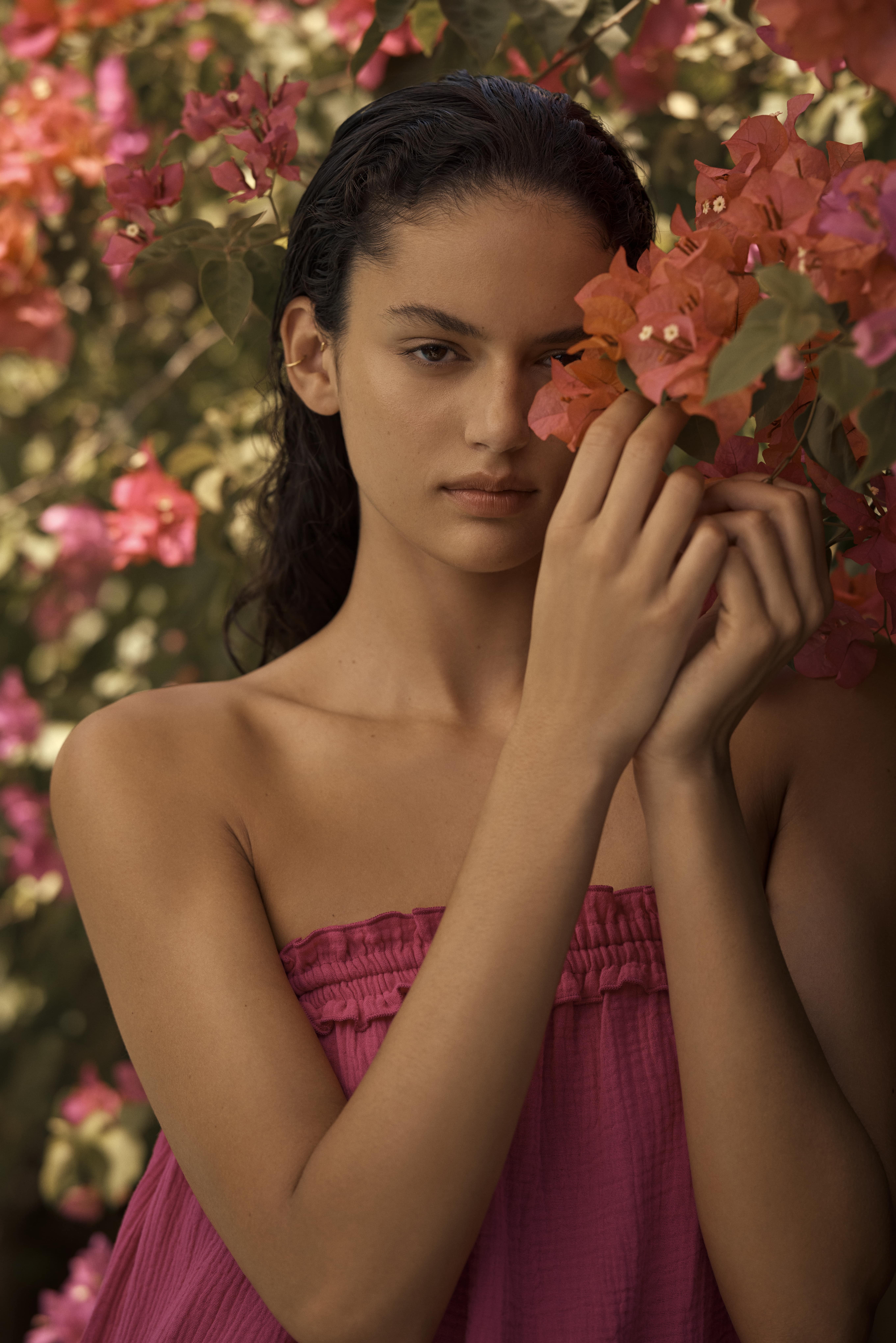 Portrait of a young woman with wet dark hair and a magenta strapless dress standing in front of a bougainvillea bush with coral flowers.