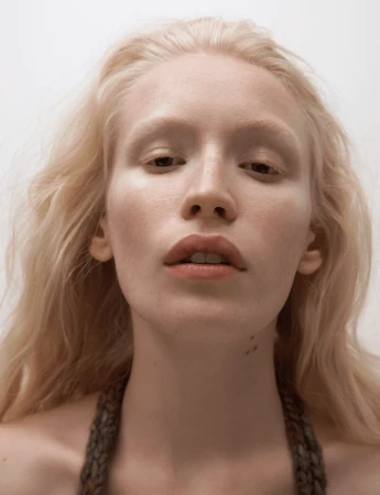 Close-up portrait of a fair-skinned model with blonde hair and natural makeup against a plain background.