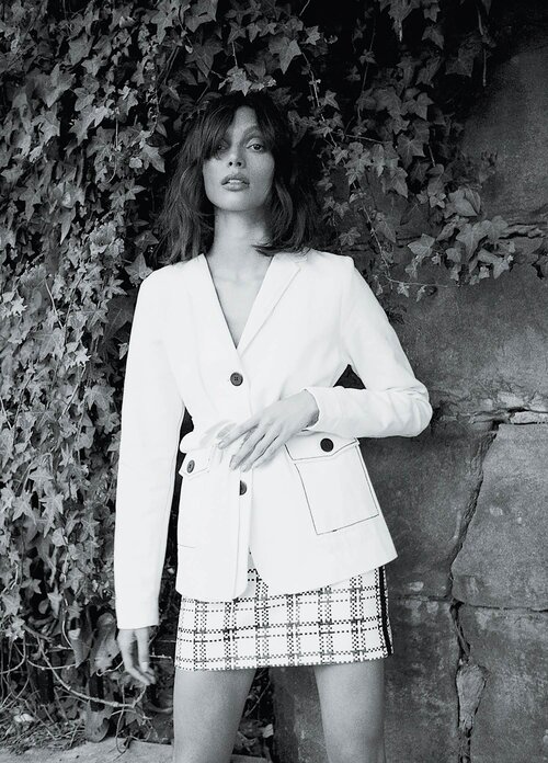 Model in white blazer and patterned skirt posing against an ivy-covered stone wall, shot in monochrome.