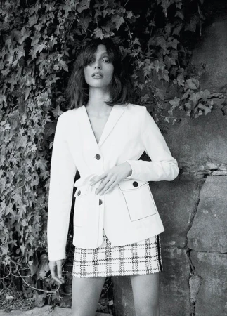Model in white blazer and patterned skirt posing against an ivy-covered stone wall, shot in monochrome.