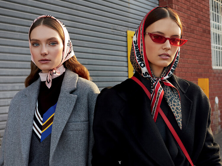 Two fashion models in headscarves and coats pose in front of a gray metal wall, one in red sunglasses, for an outdoor fashion shoot.