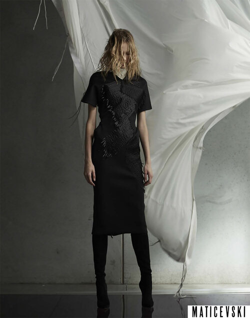 A full-length shot of a model in a black dress with an asymmetrical design and thigh-high boots, against a neutral gray backdrop.