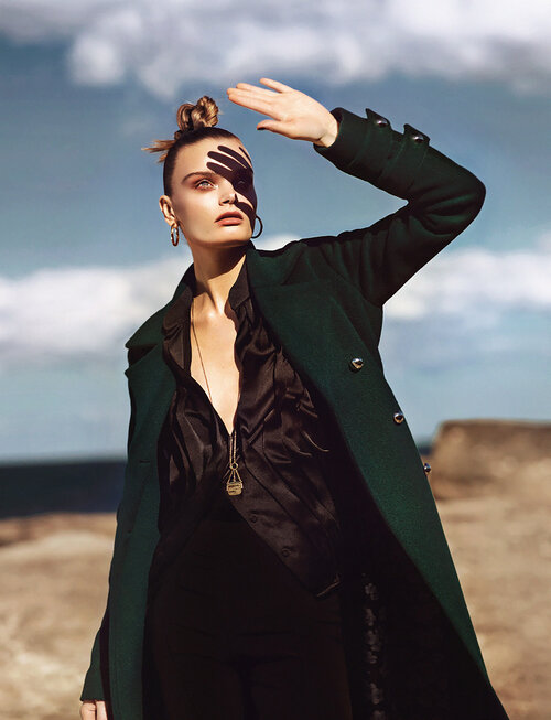 Woman in stylish green coat shields eyes from sunlight outdoors, wearing black top and pendant necklace against a sky backdrop.
