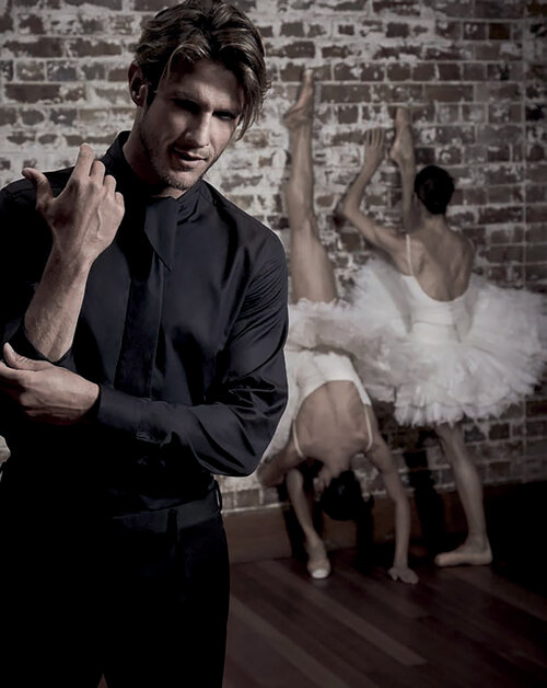 Stylish male model in a black shirt poses as three ballerinas in white tutus playfully stretch against a brick wall in the background.