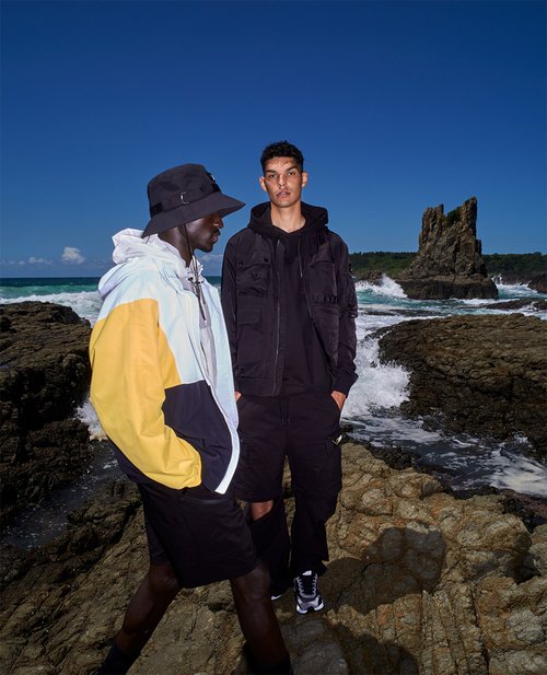 Two adult men on a rocky beach. One man wears a color-blocked jacket and bucket hat. The other wears a black jacket and shorts.