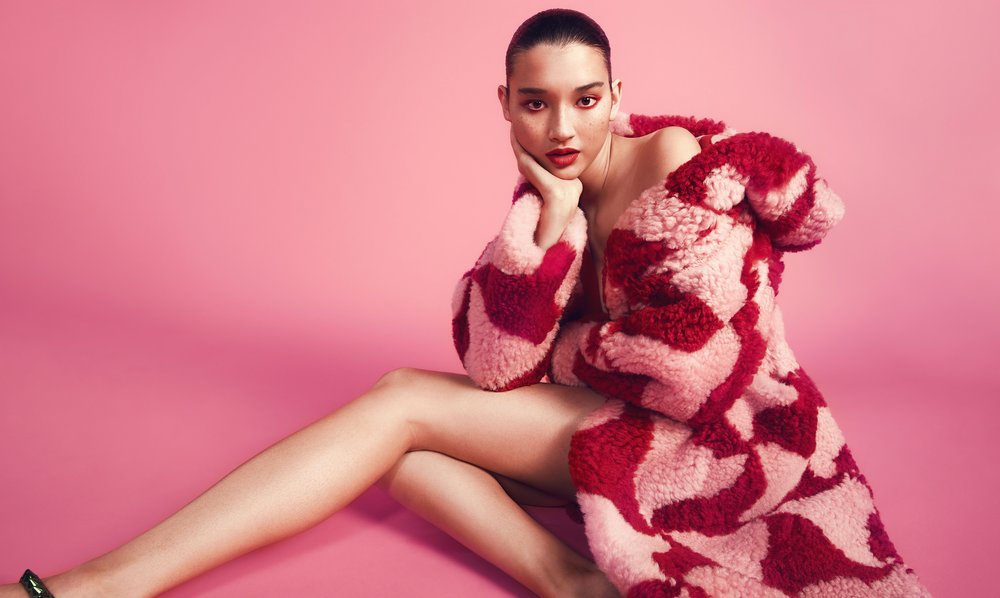 Woman in a pink and red coat sits on a pink background with her hand on her face looking at the camera in a fashion studio shot.
