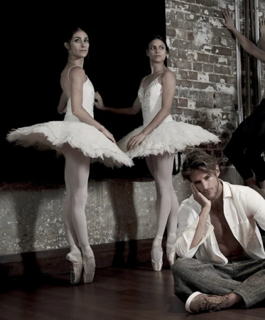 Two ballerinas in white tutus stand near a man in a white shirt seated on the floor in a dance studio with brick walls.