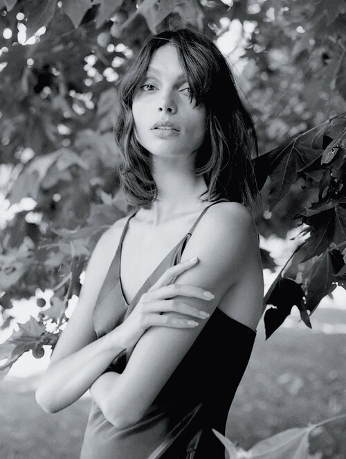 Monochrome portrait of a woman with shoulder-length hair, wearing a slip dress, standing in front of leaves.