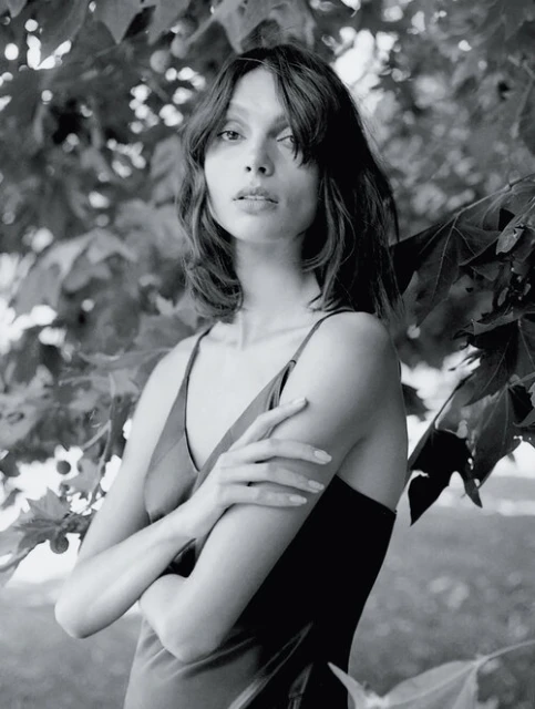 Monochrome portrait of a woman with shoulder-length hair, wearing a slip dress, standing in front of leaves.