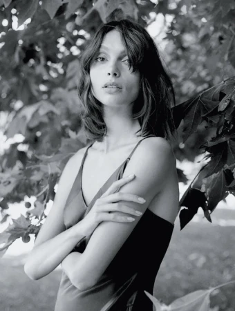 Monochrome portrait of a woman with shoulder-length hair, wearing a slip dress, standing in front of leaves.