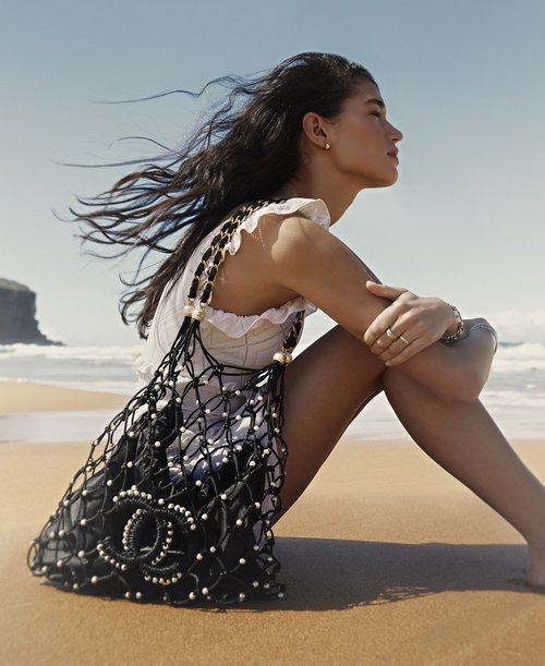 Brunette woman in a white top sits on a sandy beach with a Chanel handbag, enjoying a sunny day with a blue sky and ocean backdrop.