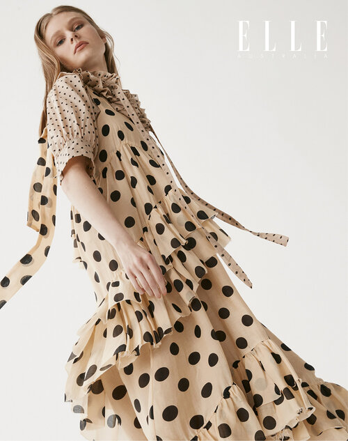 A slender woman is featured in a studio shot, wearing a light beige, tiered, polka-dot dress with a flowing tie collar.