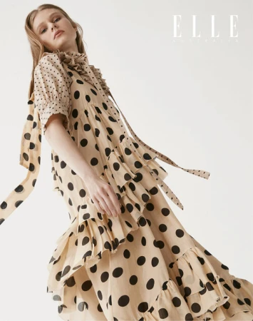 A slender woman is featured in a studio shot, wearing a light beige, tiered, polka-dot dress with a flowing tie collar.