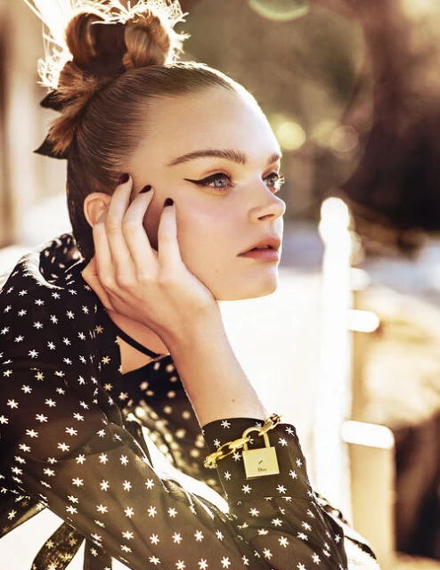 Portrait of a fair-skinned woman with winged eyeliner and a patterned blouse resting her face on her hand in an outdoor setting.