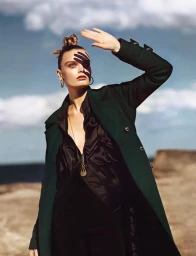 Model in dark green coat and black blouse shielding eyes under a cloudy sky in Sydney, hair styled by Keiren Street