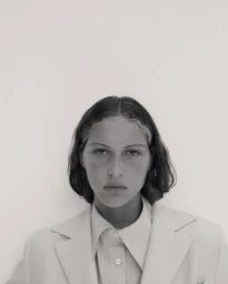 Portrait of a model with dark hair wearing a light-coloured collared shirt and blazer against a plain background
