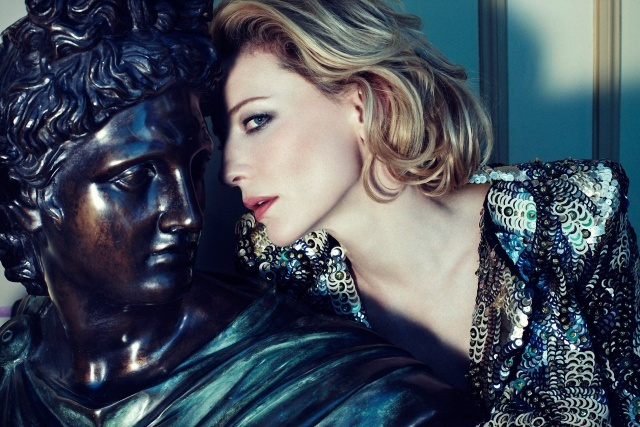 A woman with blonde hair poses in front of a bust, wearing a shiny, scale-like top in a portrait-style shot. Fashion and beauty theme.