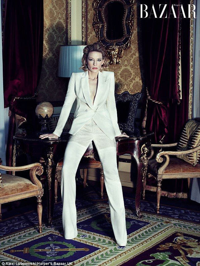 A woman with short blonde hair poses in a white suit and pants in an ornately decorated interior with a table and chairs.