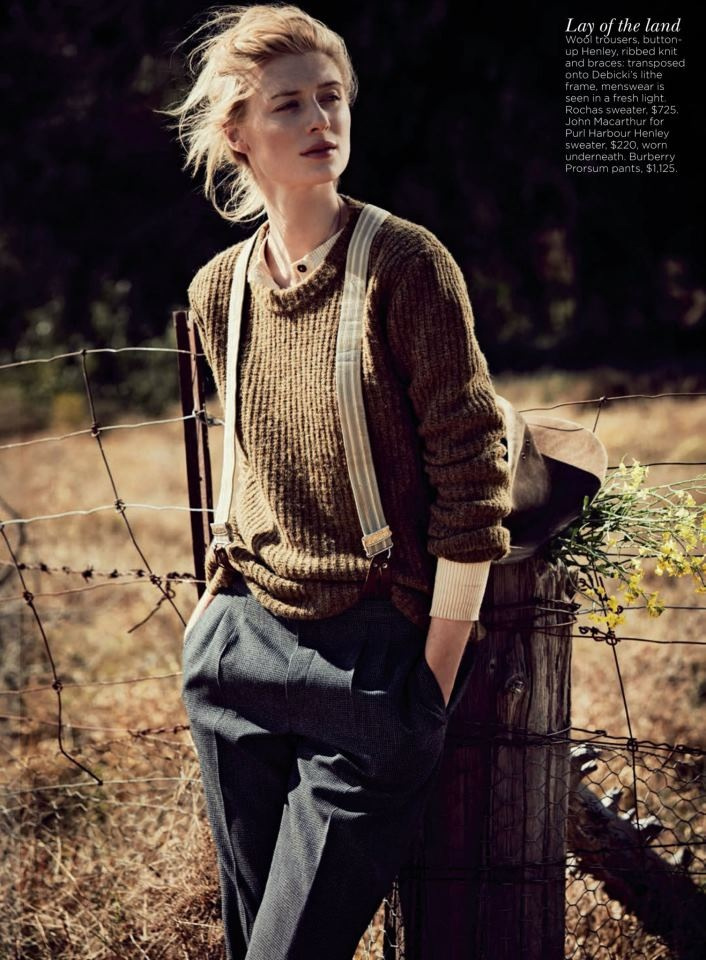 Woman in brown sweater and suspenders poses outdoors near a fence. The setting includes a rural background with earth tones.