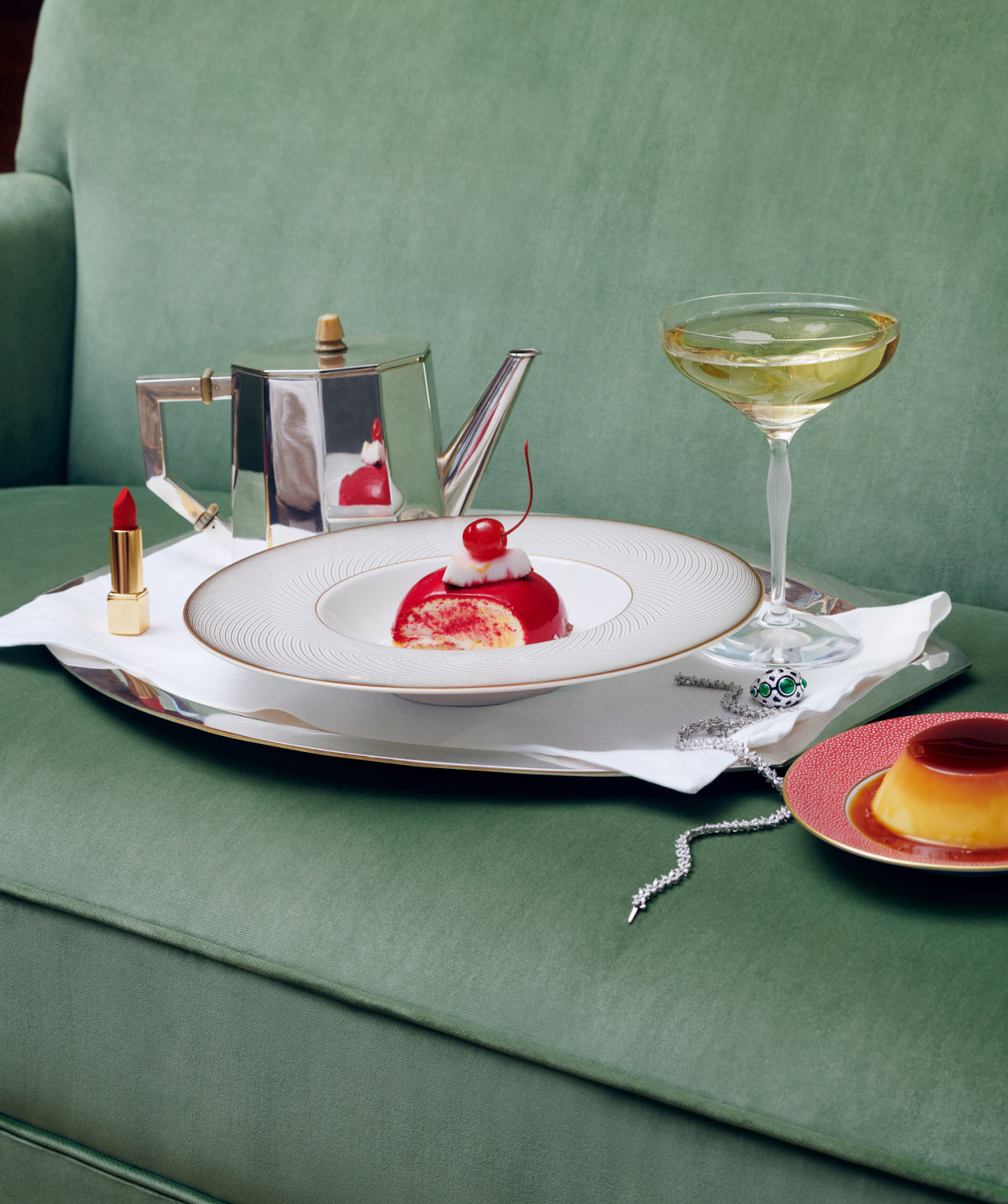 Still life composition with a dessert topped with a cherry, a teapot, champagne glass, lipstick, and jewelry on a green couch.