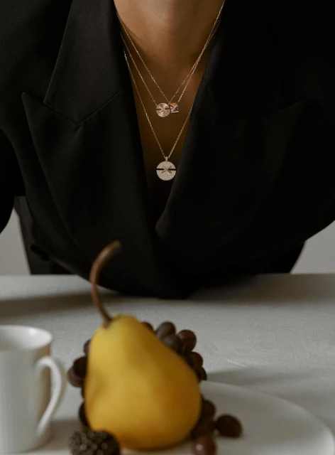 Close-up of a person wearing layered gold necklaces, complemented by a pear, grapes, and coffee cup on a neutral surface.