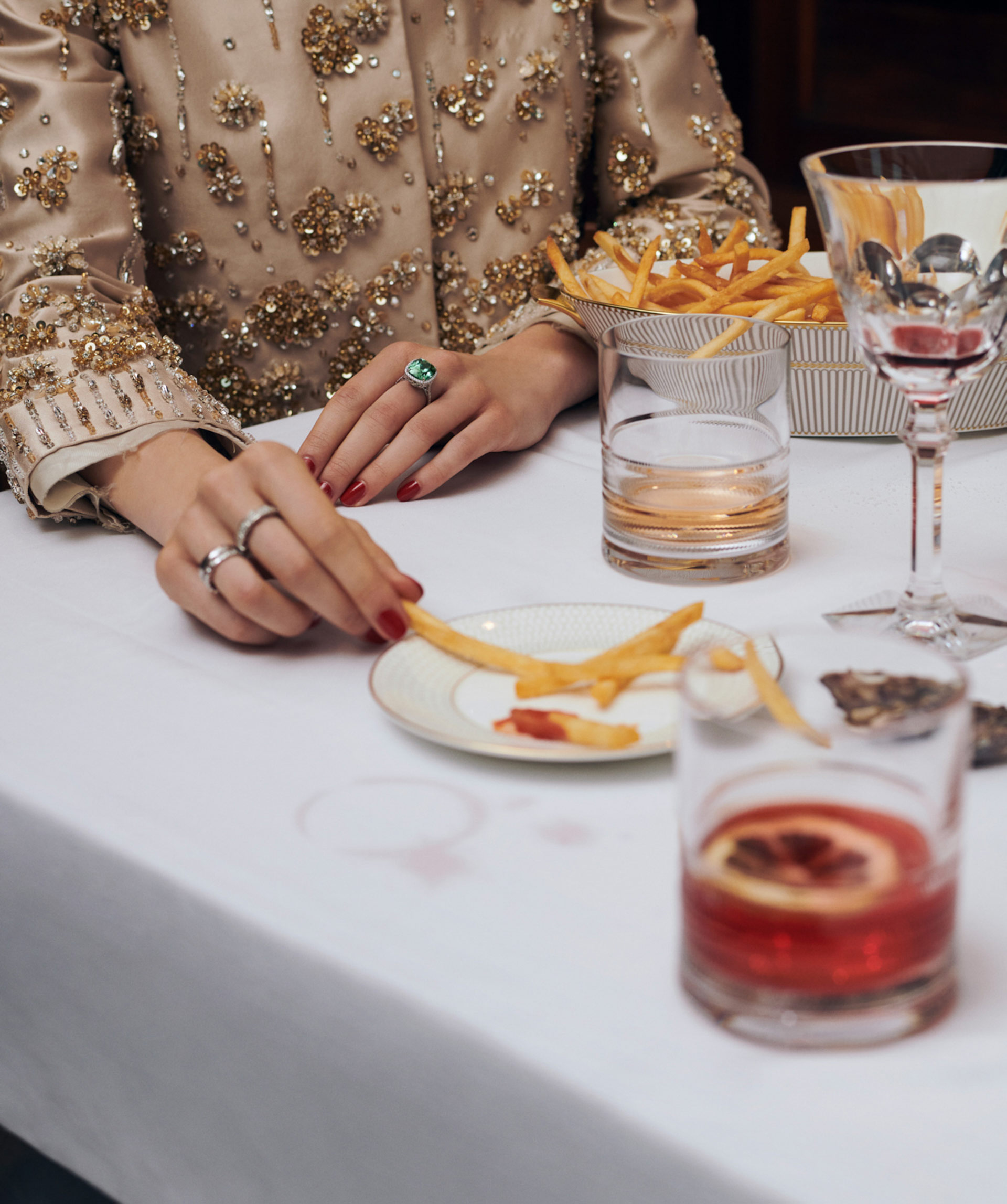 Elegant lifestyle still life: woman's hands, jewelry, and French fries at a table with drinks, illuminated by soft, warm lighting.