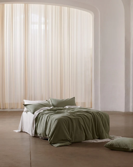 Bedroom interior with an unmade bed featuring green linen bedding, set against a backdrop of soft, natural light filtering through sheer curtains.