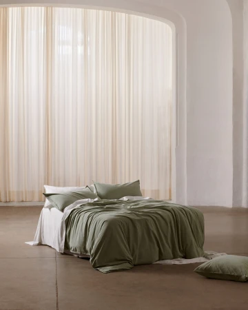 Bedroom interior with an unmade bed featuring green linen bedding, set against a backdrop of soft, natural light filtering through sheer curtains.