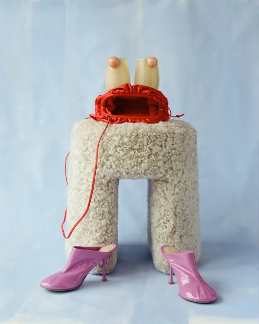 Still life of designer fashion accessories on a pale blue background: fluffy stool, open red purse, lucite earrings, and pink heeled mules.