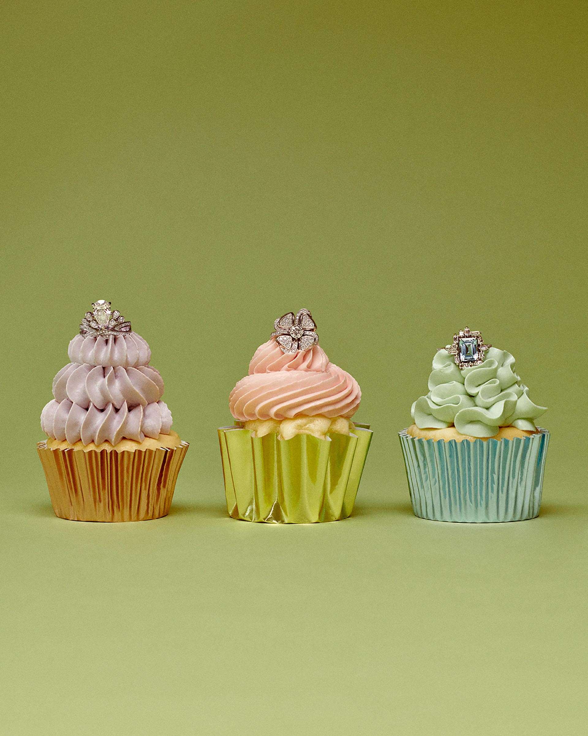 Three cupcakes with pastel frosting and rings on top, displayed in a row against a light green background in a studio setting.