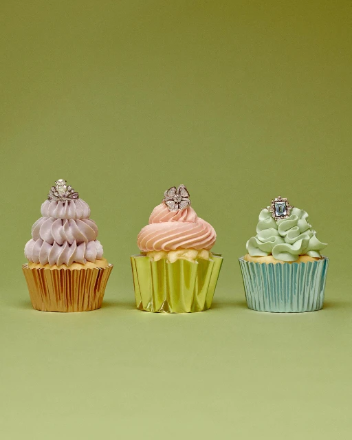 Three cupcakes with pastel frosting and rings on top, displayed in a row against a light green background in a studio setting.