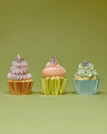 Three cupcakes with pastel frosting and rings on top, displayed in a row against a light green background in a studio setting.