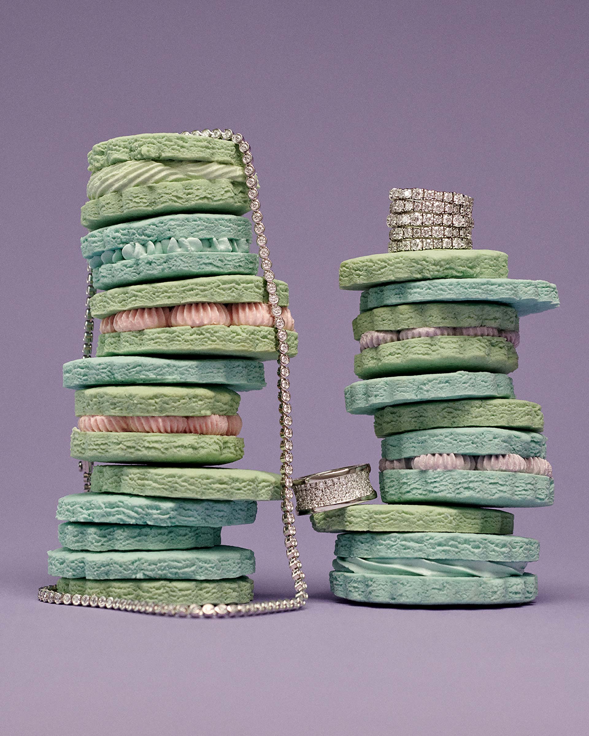 Stack of pastel coloured sponge cakes with diamond jewellery accent on a light purple background in a studio setting.
