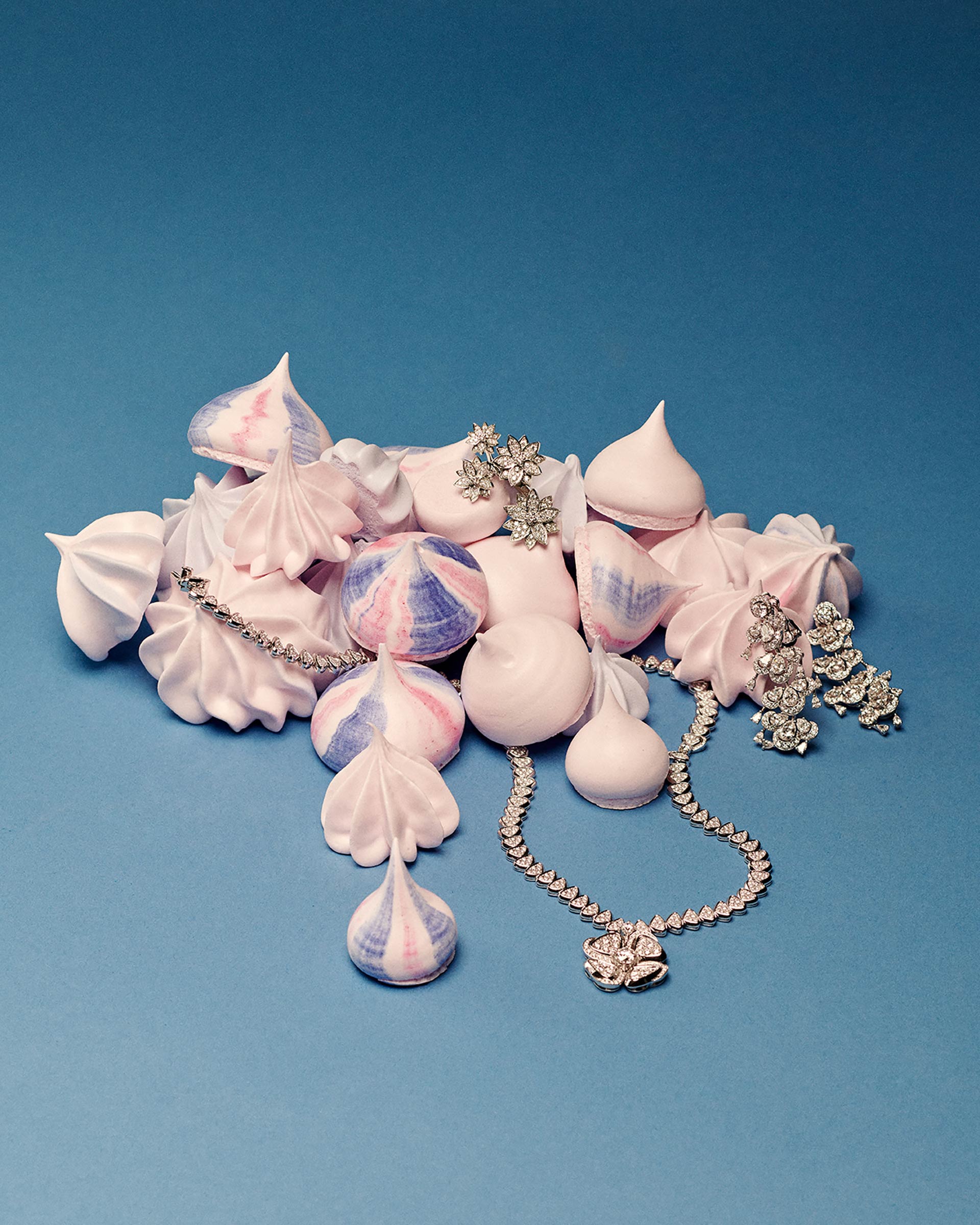 Elegant still life photo features pastel meringues, a diamond necklace, earrings, and brooch against a light blue background.