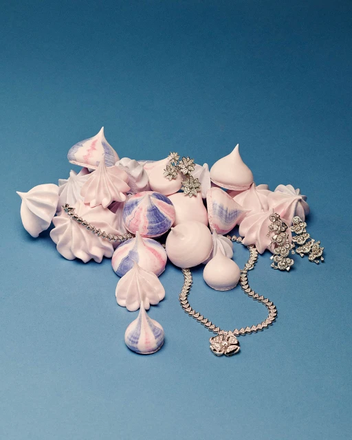 Elegant still life photo features pastel meringues, a diamond necklace, earrings, and brooch against a light blue background.