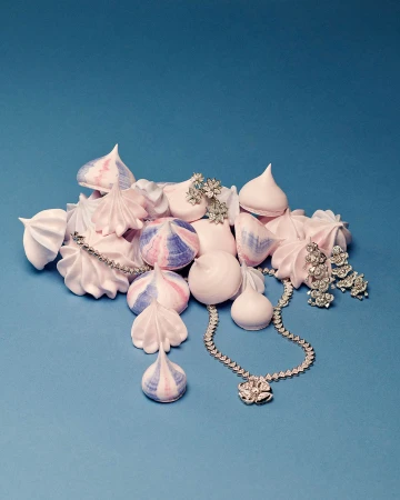 Elegant still life photo features pastel meringues, a diamond necklace, earrings, and brooch against a light blue background.