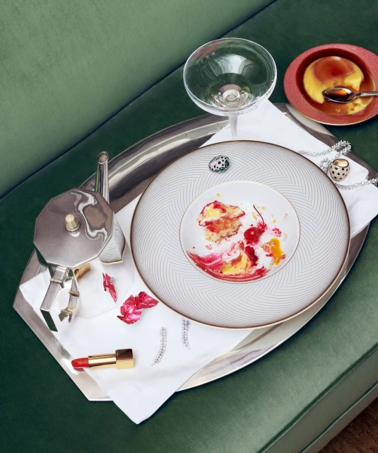 Overhead shot of a silver tray with a dessert plate, cocktail glass, coffee maker, jewelry, and lipstick on a green upholstered surface.