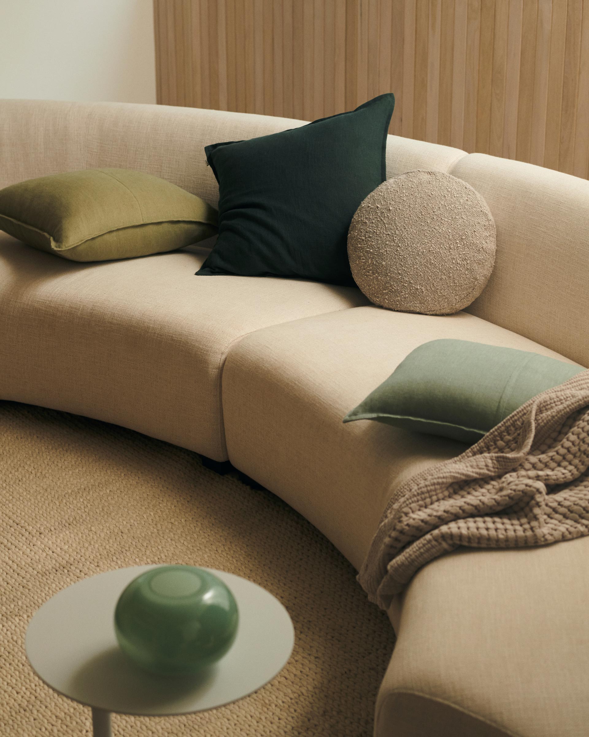 Beige curved sofa with green and beige cushions, a small white table with a green glass ornament and a beige carpet.