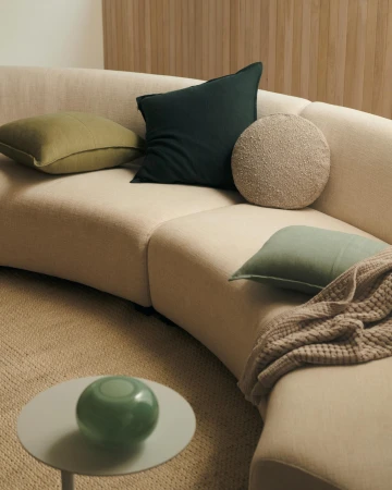 Beige curved sofa with green and beige cushions, a small white table with a green glass ornament and a beige carpet.