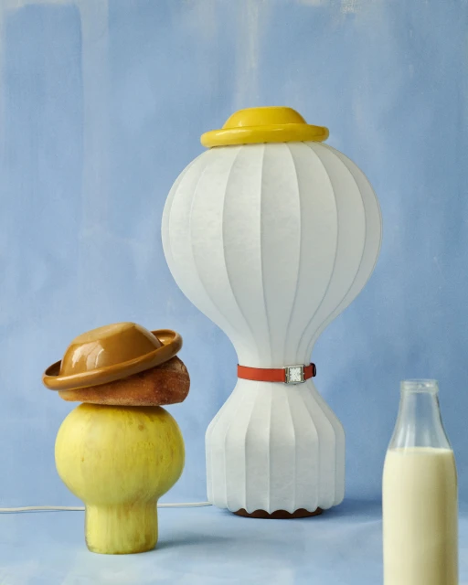 Still life featuring a lamp with a yellow hat, a yellow vase with a brown hat and bread, and a bottle of milk against a blue backdrop.