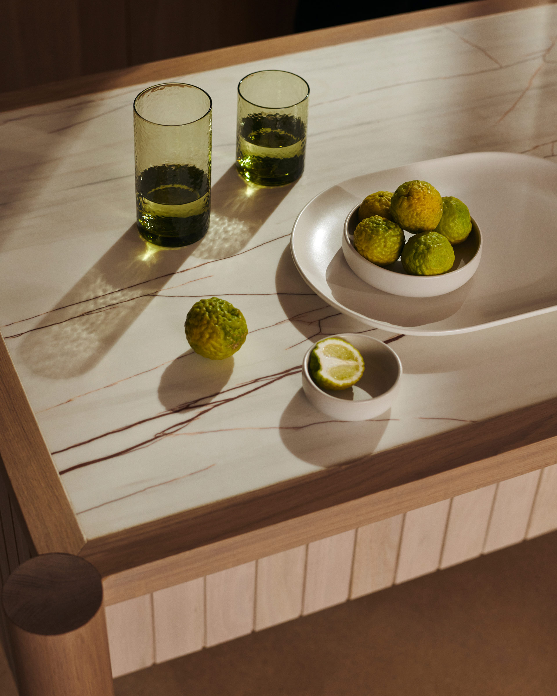 Still life photo of fresh citrus fruits and glasses with liquid on a marble-topped wooden table, illuminated by natural light.