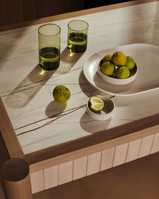 Still life photo of fresh citrus fruits and glasses with liquid on a marble-topped wooden table, illuminated by natural light.