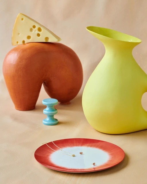 Still life shot of a triangular slice of cheese on an orange vase, near a yellow vase, a blue spool, and a white and red plate.